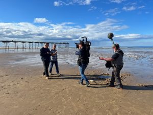 Actors Mark Benton and Robson Green walking along Saltburn beach eating an ice cream being filmed by a camerman with a soundman behind them