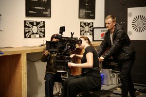 2 ladies looking through a TV camera lens being pushing along a track by a man