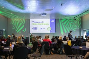 People around round tables at an event looking at a large screen on the wall